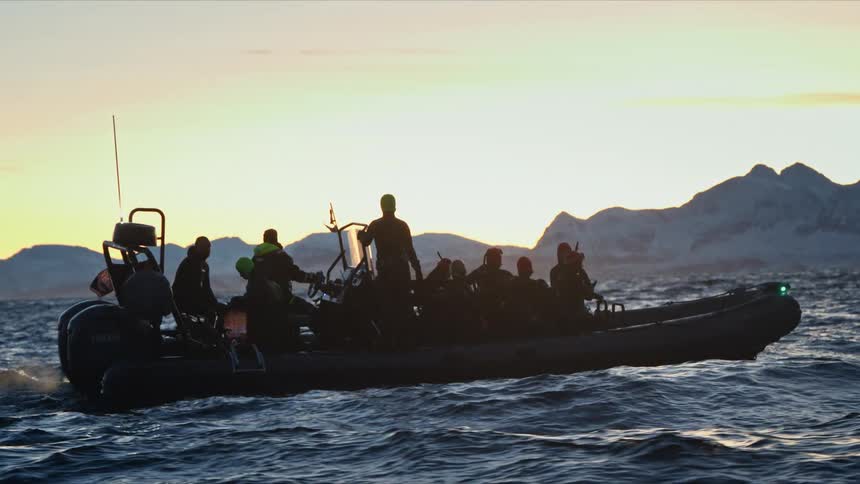 A silhouetted group of adventurers on a rigid inflatable boat tour floats on the arctic sea at sunset with snowy mountains in the background