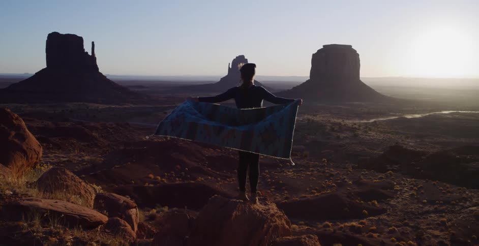A silhouetted woman holds a colorful patterned rug open while standing on a red rock overlooking the iconic buttes of Monument Valley at sunrise