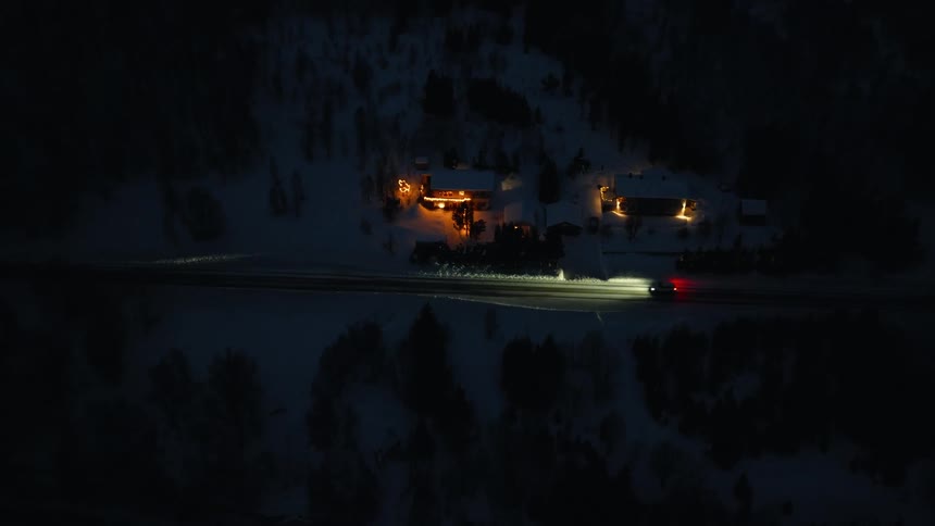 An aerial view of a car driving on a remote road at night, passing by warmly lit houses in a snowy winter landscape