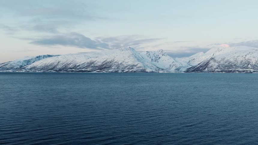 A majestic range of snow-covered mountains rises above the calm, cold waters of a Norwegian fjord
