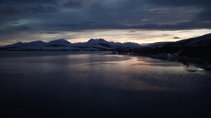 An aerial shot flies over a car-filled bridge and a dark, icy fjord at twilight, with the last light of sunset glowing behind snow-covered mountains