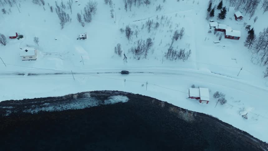 An aerial top-down view shows a car driving on a snow-covered road along the coast of a remote Nordic village