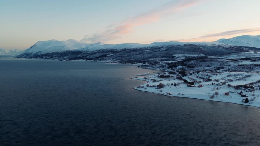 A static aerial view captures a quiet, snow-covered village on the edge of a fjord in Norway during a beautiful winter sunset