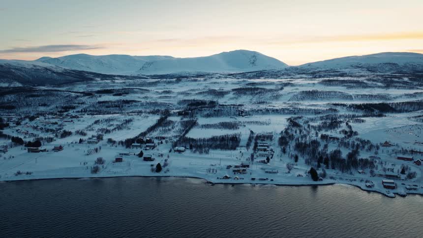 A stunning aerial view of a remote village on the shore of a fjord, surrounded by a vast snowy landscape and mountains during a winter sunset