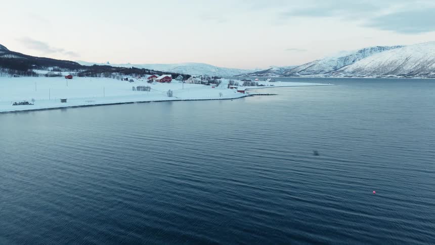 A serene aerial shot glides over a rippling blue fjord towards a snow-covered village nestled at the foot of towering winter mountains in Norway