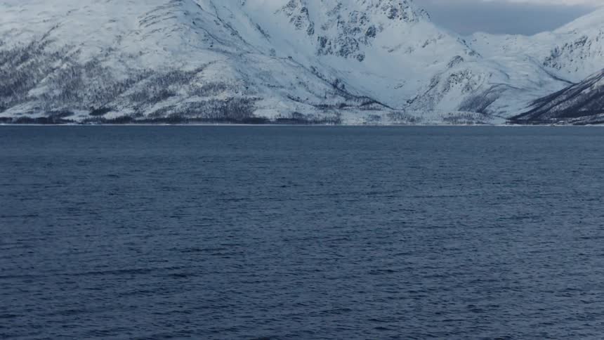 A slow tilt up from the dark, cold water of a fjord reveals a majestic, snow-covered mountain range in a vast Nordic winter landscape