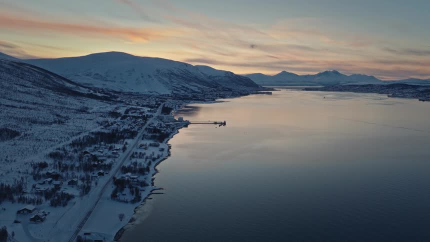 A serene aerial pullback shot reveals a snow-covered coastal village alongside a tranquil fjord during a beautiful winter sunset in Norway
