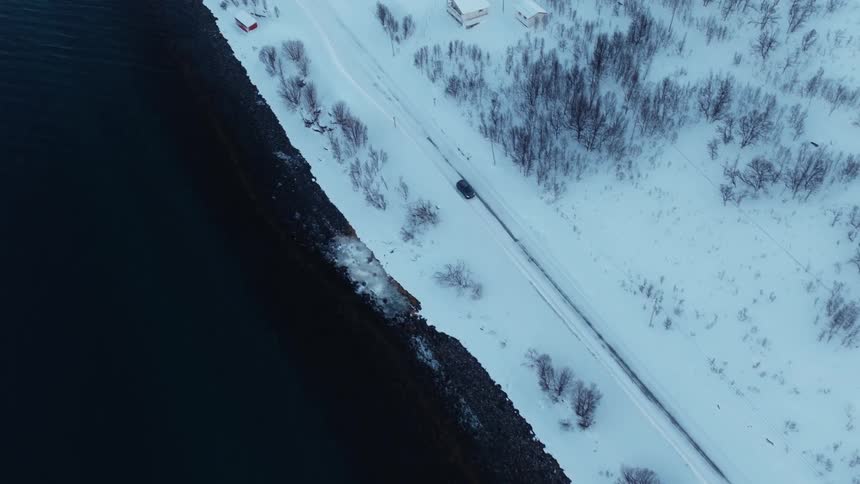 Aerial top-down shot of a car driving along a snow-covered road next to a dark fjord in a remote winter landscape