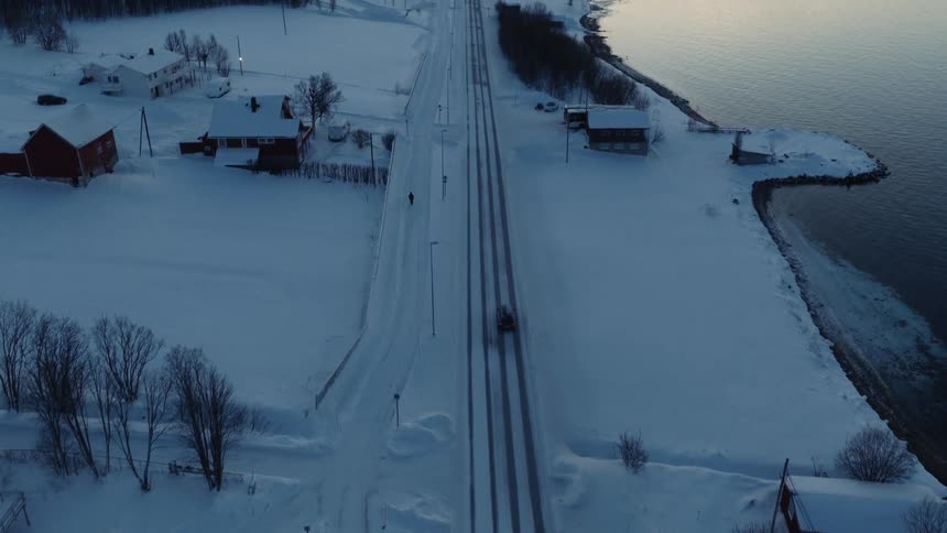 Aerial reveal shot following a vehicle on a snowy road through a coastal village nestled between a fjord and mountains at sunset