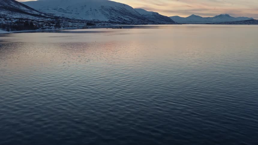 Aerial tilt-up from the rippling fjord water to reveal a stunning snowy mountain landscape at sunset
