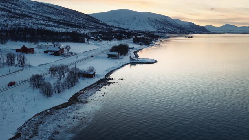 Aerial view of a tranquil fjord and a snow-covered coastal village with cars driving on the road during a winter sunset in Norway