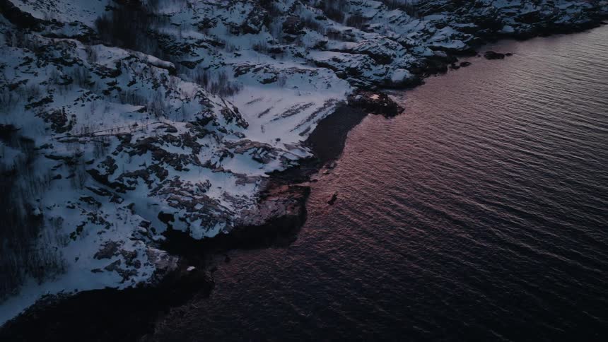 An aerial view shows a snowy, rocky coastline meeting the calm, rippling water reflecting a beautiful winter sunset