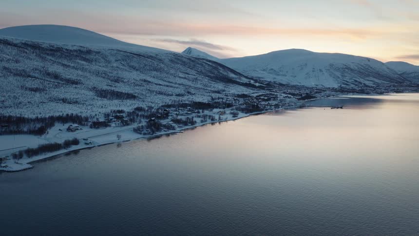 An aerial view captures the serene beauty of a remote coastal village nestled against snow-covered mountains beside a calm fjord at sunset