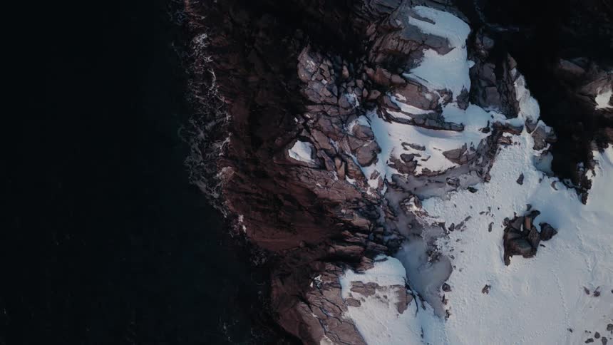 A slow-moving top-down aerial shot reveals the beautiful and stark contrast between a snowy, rocky shoreline and the dark, cold ocean