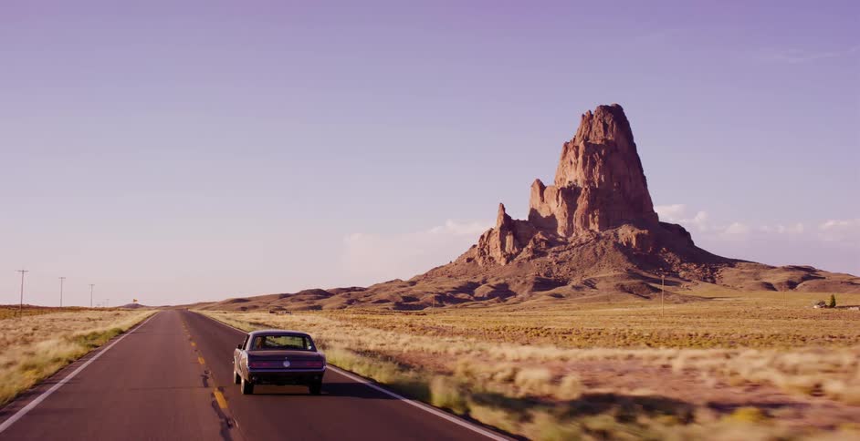 A classic car drives along a long, straight desert road towards a towering rock formation in the American Southwest