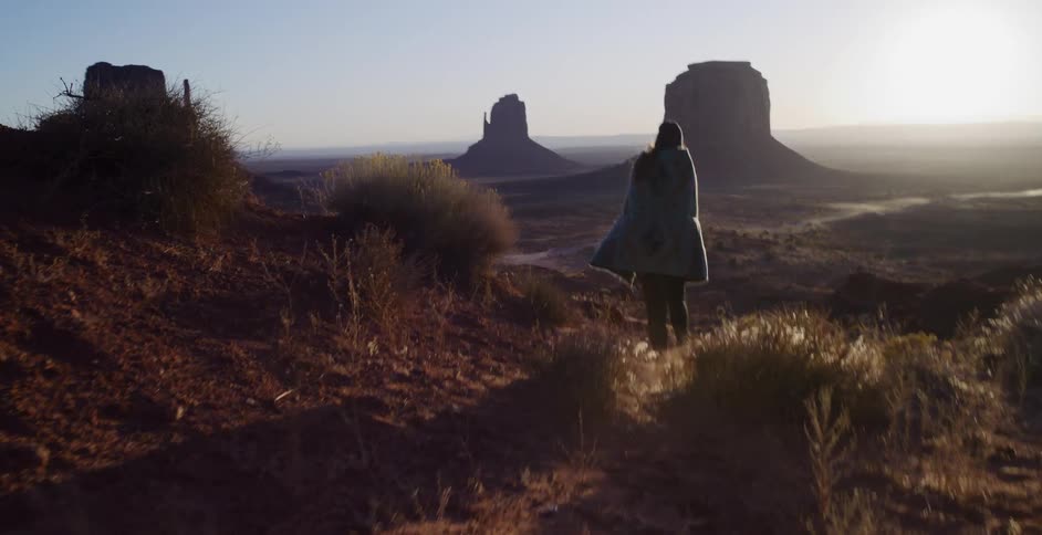 A woman wrapped in a traditional blanket walks along a dusty path overlooking the majestic buttes of Monument Valley at sunrise