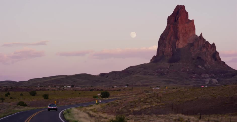 A lone car drives on a winding desert highway towards a majestic mountain peak under a full moon at dusk
