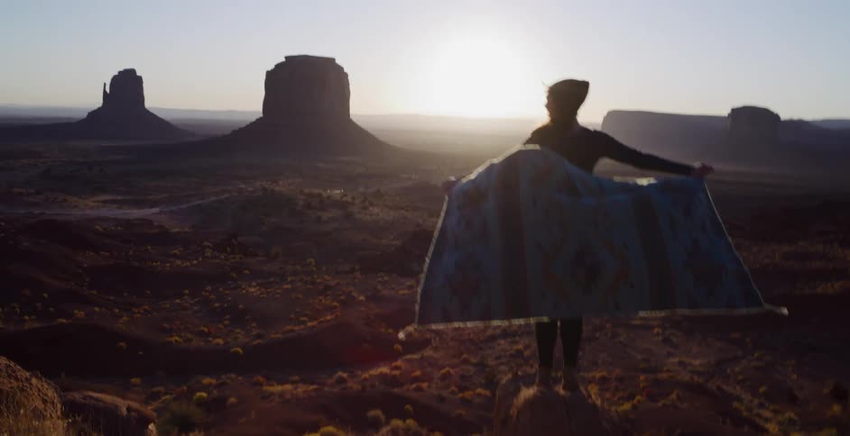 A woman holds a blanket in the wind while standing on a rock overlooking Monument Valley at sunrise