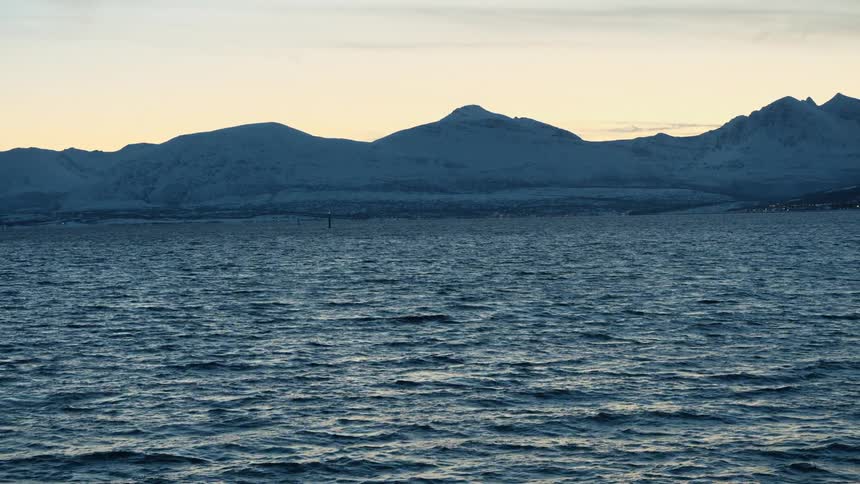 Calm ocean waves ripple in front of a majestic, snow-covered mountain range during a serene winter sunset