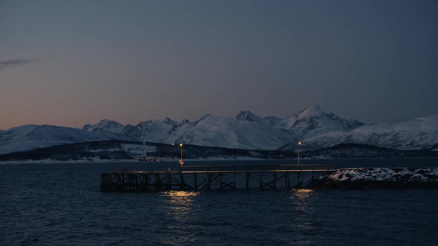 A tranquil winter twilight scene shows a lit pier on a calm fjord with a backdrop of majestic snow-covered mountains