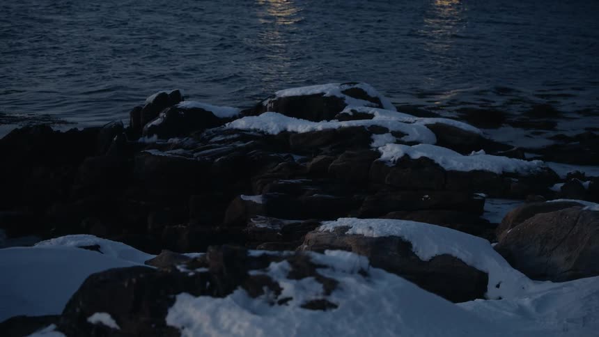 Snow-covered rocks line a dark, cold shoreline at dusk as distant lights reflect on the water's surface