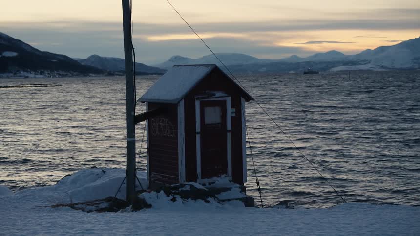 A small red hut sits on a snowy shoreline as a vast fjord and mountains fill the background at dusk