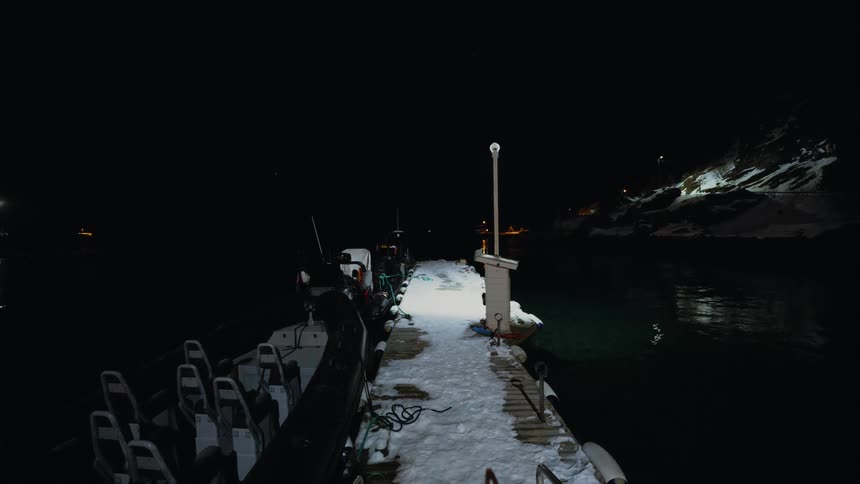 A rigid inflatable boat is docked at a snowy pier in a quiet harbor on a dark winter night