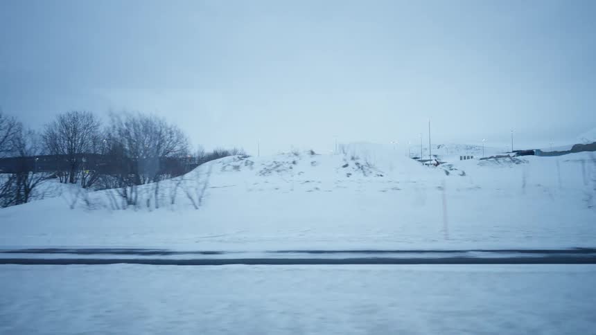 A slow-motion, side-view tracking shot from a moving vehicle reveals a cold, snow-covered landscape on an overcast winter day