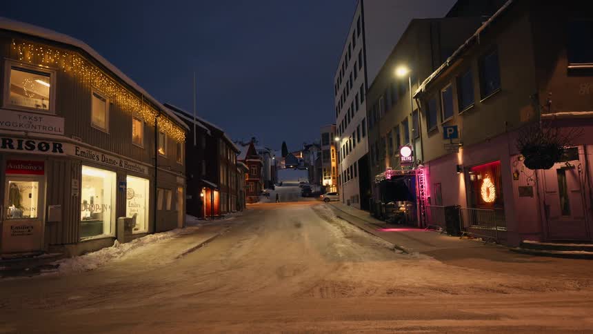 A quiet snow-covered street in a small Norwegian town glows with the warm light of storefronts and festive decorations on a winter night