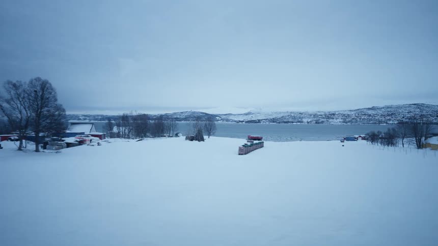 Tracking shot reveals a quiet, snowy village with cozy houses nestled beside a tranquil fjord on a cold winter day in Norway