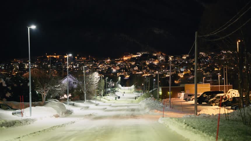 A quiet, snowy street in a northern town glows under streetlights at night, with a hillside city visible in the background