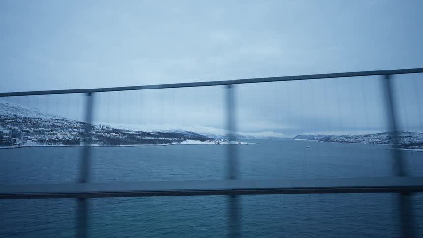 A scenic point-of-view shot from a vehicle driving across a bridge overlooking a vast fjord and a snowy coastal town in winter