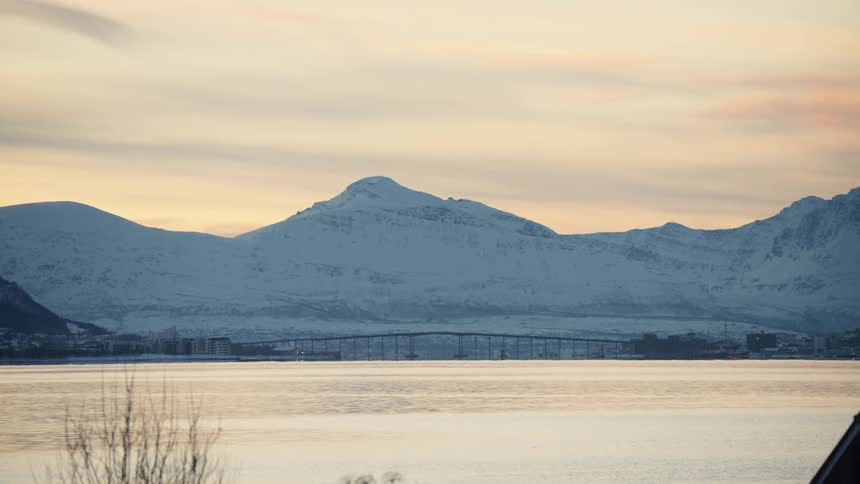 A tranquil view of a bridge crossing a fjord with a majestic, snow-covered mountain range illuminated by the soft light of a winter sunset