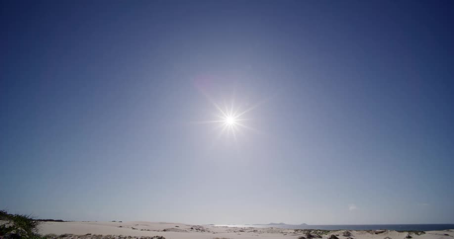 The bright midday sun shines in a clear, cloudless blue sky over a vast desert landscape of sand dunes next to the ocean