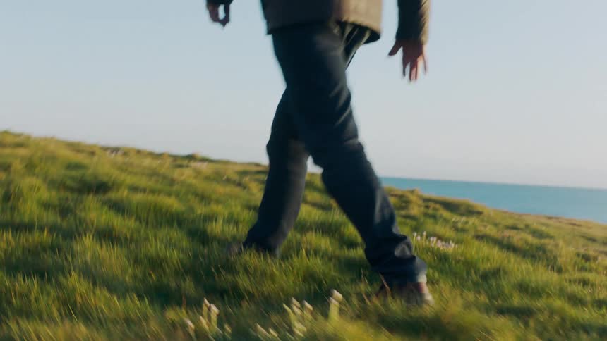 A close-up slow-motion shot follows a man's legs and boots as he walks through a grassy field towards the ocean
