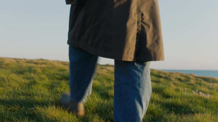 Low-angle slow motion shot follows a person's boots walking through a grassy field overlooking the ocean