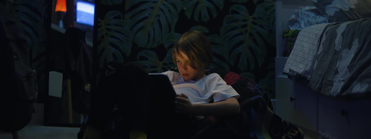 A young boy sits on the floor of his dimly lit bedroom at night, captivated by the glow of his tablet as the camera slowly zooms in