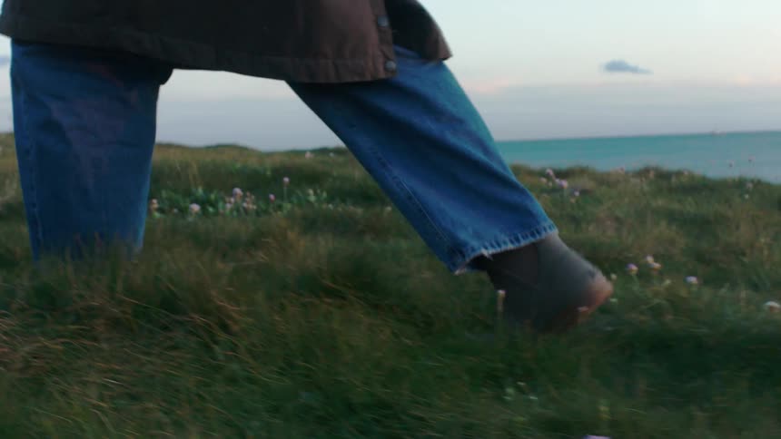 A person in boots and jeans walks in slow motion through a grassy, flower-strewn cliffside overlooking the ocean at dusk