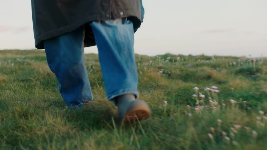 A low-angle shot of two people's feet in boots walking through a grassy, flower-filled meadow along a coastal cliff at dusk