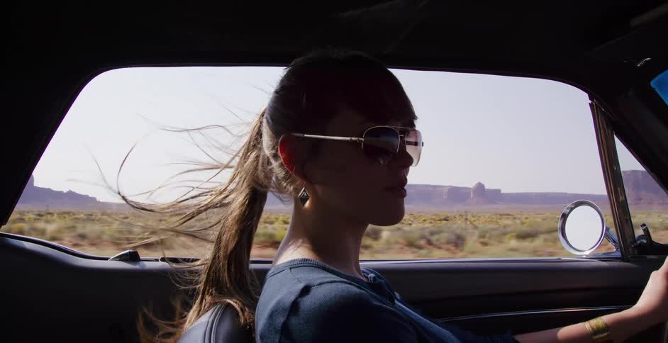 A woman with sunglasses drives a vintage convertible through the desert as the wind blows through her hair in slow motion