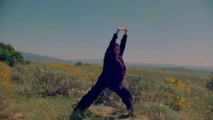 A woman with red hair performs slow, graceful yoga stretches in a sun-drenched field of yellow wildflowers
