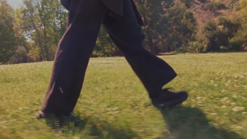 A low-angle tracking shot follows a person's legs as they walk through a sunny, green meadow dotted with white flowers
