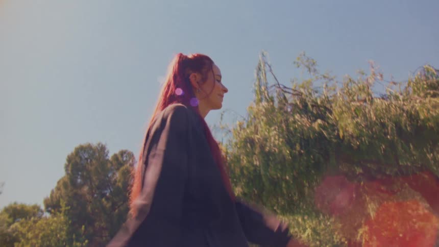 A woman with vibrant red hair walks in slow motion through a sunlit park, filmed from a low angle with dreamy lens flares