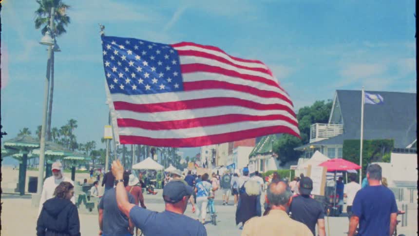 Vintage-style shot of a crowd walking along a sunny beach boardwalk as a man holds up an American flag