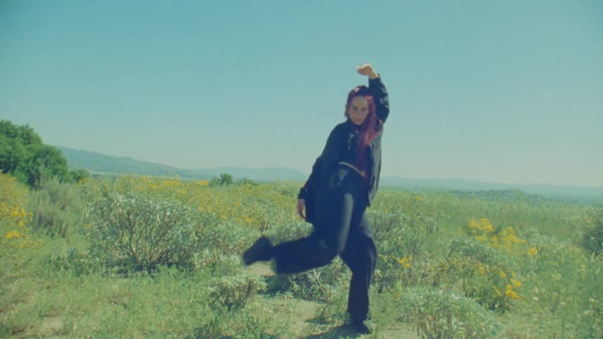 A woman with red hair performs an expressive dance in a sunny wildflower field, filmed with a vintage aesthetic