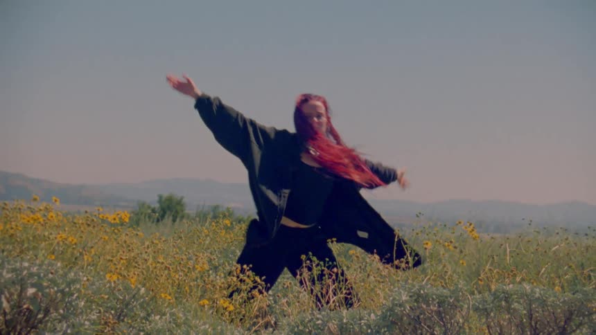 A woman with vibrant red hair performs an expressive, slow-motion dance in a sun-drenched field of yellow wildflowers