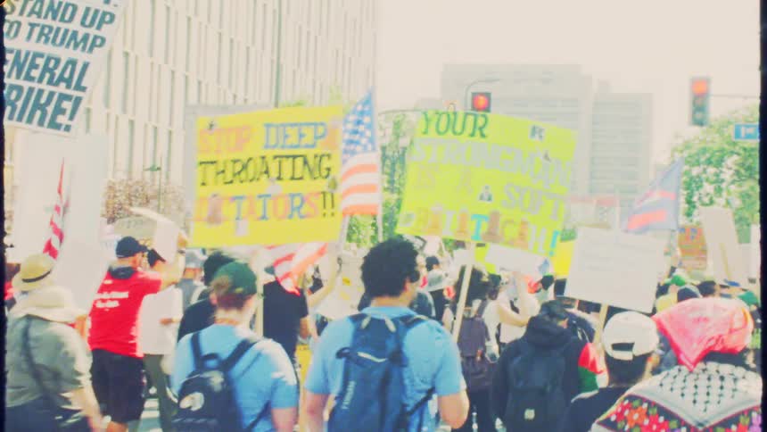 Vintage handheld footage captures a large crowd of protestors marching with political signs down a sunny city street