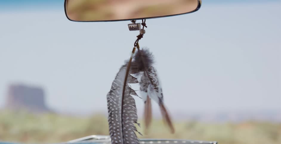 A slow-motion close-up of decorative feathers hanging from a car's rearview mirror as they gently sway on a desert road trip