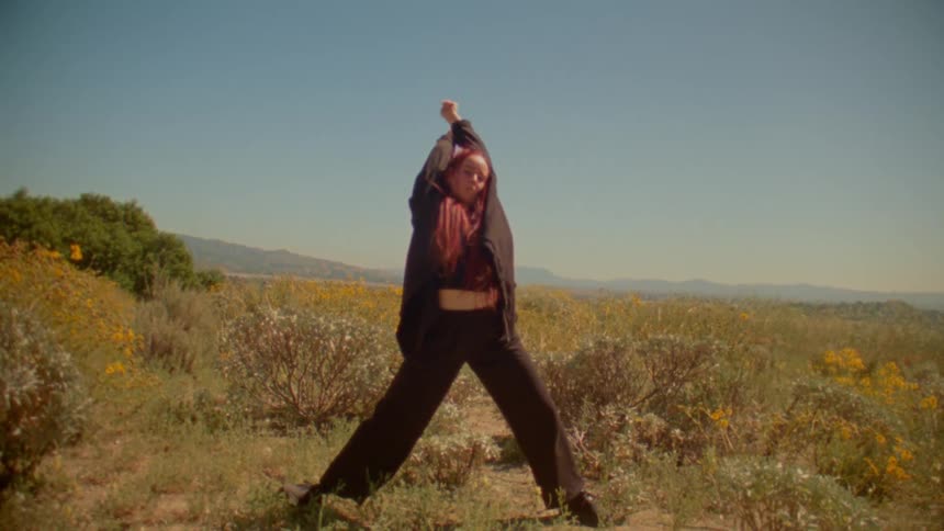 A person with long red hair performs a slow, expressive dance in a wide-open, sunlit field