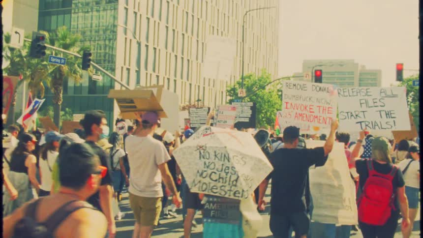 A vintage-style slow-motion shot follows a crowd of protestors marching down a city street with signs and flags